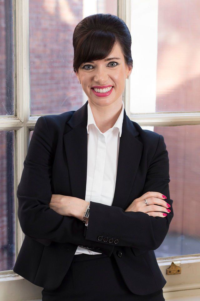 A woman dressed in a professional black suit is standing with her arms crossed and smiling in front of a window.