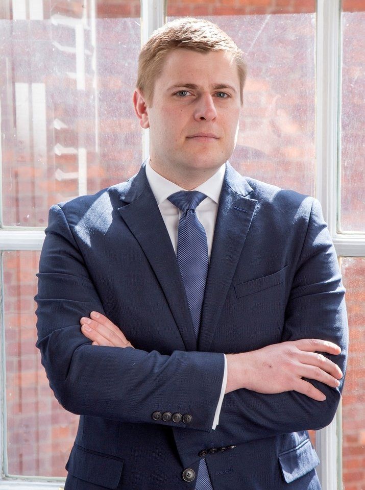 A man in a blue suit and tie stands with his arms crossed in front of a large window.